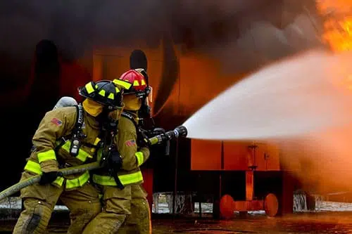 "Ein Feuerwehrmann in Warnschutzkleidung steht neben einem Feuerwehrfahrzeug auf einer asphaltierten Straße. Im Hintergrund sind Gasflammen zu sehen, während der Feuerwehrmann einen Helm trägt und sich auf den Einsatz vorbere